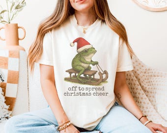 May include: Cream-colored t-shirt featuring a green frog wearing a Santa hat and riding a wooden sled. The text "off to spread christmas cheer" is printed below the frog. The shirt is a festive holiday design.