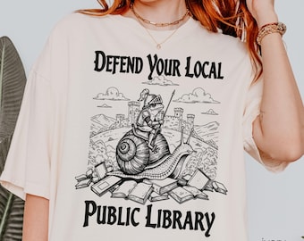 May include: An ivory t-shirt featuring a black graphic print of a frog wearing knight armor and riding a snail in front of a castle. The text reads DEFEND YOUR LOCAL PUBLIC LIBRARY. The design includes scattered books at the base of the illustration.