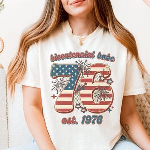 May include: Cream-colored t-shirt featuring a patriotic design. The graphic includes the text "bicentennial babe" above a stylized "76" with a red and white striped pattern and a blue star field. The text "est. 1976" is below.