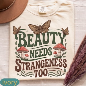 Może przedstawiać: Kremowa koszulka z napisem "BEAUTY NEEDS STRANGENESS TOO" w kolorze zielonym i brązowym. Projekt zawiera motyle i grzyby z czerwonymi kapeluszami. Rozmiar XL.
