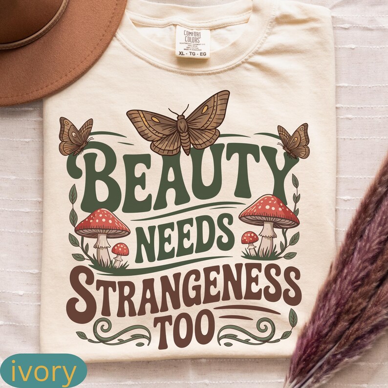 Może przedstawiać: Kremowa koszulka z napisem "BEAUTY NEEDS STRANGENESS TOO" w kolorze zielonym i brązowym. Projekt zawiera motyle i grzyby z czerwonymi kapeluszami. Rozmiar XL.