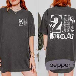May include: A charcoal gray t-shirt featuring a white graphic print. The front displays a small number 2 and a shot glass. The back features a large number 2, a stack of three shot glasses, and the text 21st BIRTHDAY with stars and checkered patterns.