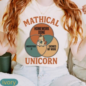 Peut inclure: T-shirt ivoire avec un motif de licorne et les mots "Mathical Unicorn". Le motif comprend des cercles qui se chevauchent avec le texte : "Homework Done", "Understood It" et "Showed My Work". La licorne tient un livre.