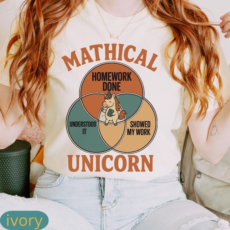 Peut inclure: T-shirt ivoire avec un motif de licorne et les mots "Mathical Unicorn". Le motif comprend des cercles qui se chevauchent avec le texte : "Homework Done", "Understood It" et "Showed My Work". La licorne tient un livre.