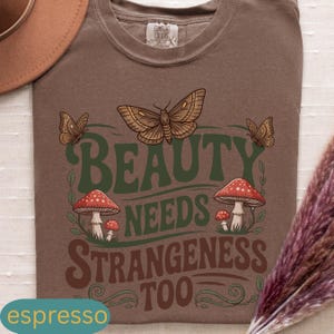 Może przedstawiać: Koszulka w kolorze espresso z graficznym wzorem przedstawiającym dużego motyla, grzyby i napis "BEAUTY NEEDS STRANGENESS TOO" w kolorze zielonym i brązowym. Koszulka ma okrągły dekolt i kr&oacute;tkie rękawy.