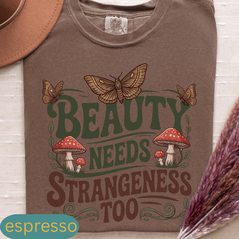 Może przedstawiać: Koszulka w kolorze espresso z graficznym wzorem przedstawiającym dużego motyla, grzyby i napis "BEAUTY NEEDS STRANGENESS TOO" w kolorze zielonym i brązowym. Koszulka ma okrągły dekolt i kr&oacute;tkie rękawy.