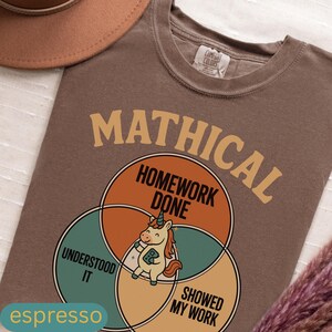 Peut inclure: T-shirt couleur expresso avec un motif graphique repr&eacute;sentant le mot "MATHICAL" au-dessus d'un diagramme de Venn. Le diagramme comprend les phrases "HOMEWORK DONE", "UNDERSTOOD IT" et "SHOWED MY WORK", avec une licorne de dessin anim&eacute; au centre.