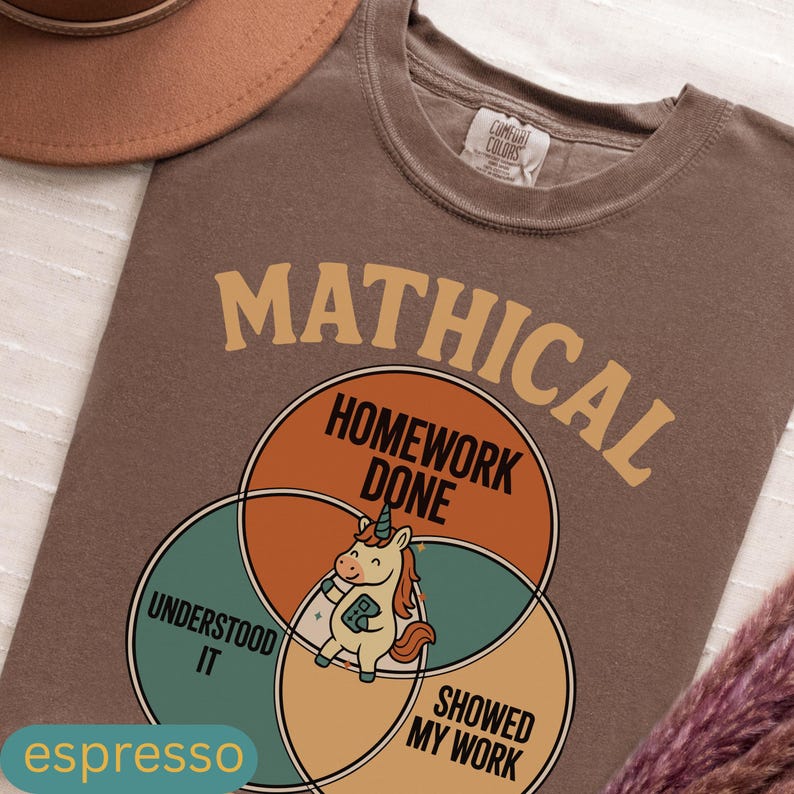 Peut inclure: T-shirt couleur expresso avec un motif graphique repr&eacute;sentant le mot "MATHICAL" au-dessus d'un diagramme de Venn. Le diagramme comprend les phrases "HOMEWORK DONE", "UNDERSTOOD IT" et "SHOWED MY WORK", avec une licorne de dessin anim&eacute; au centre.