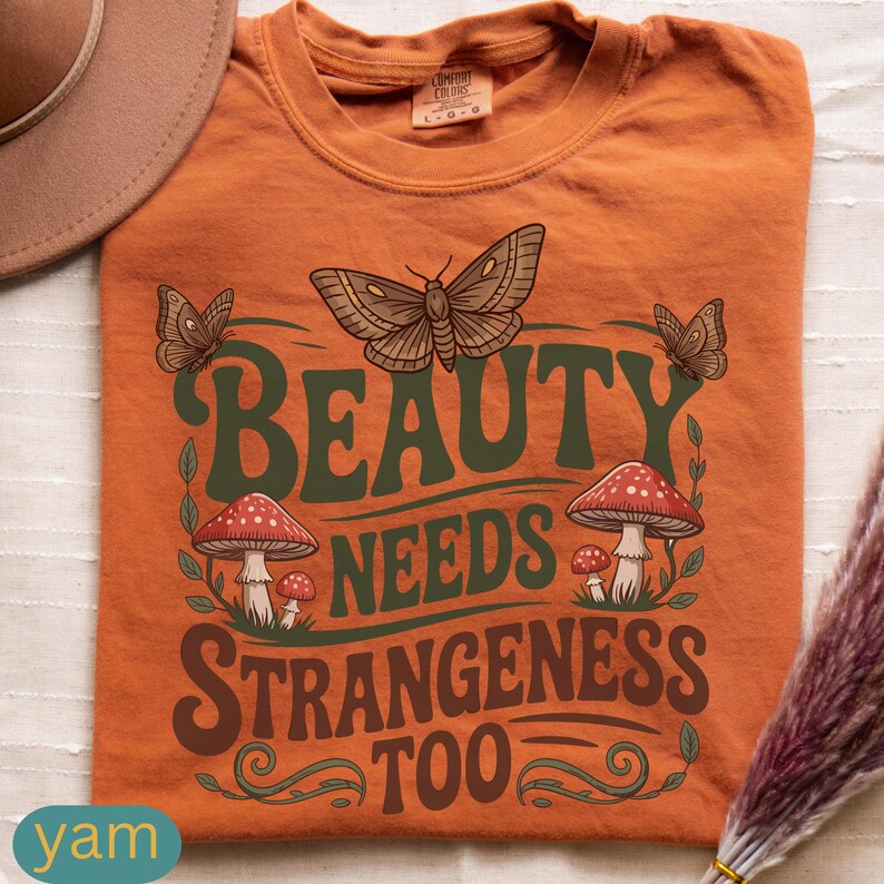 Może przedstawiać: Pomarańczowa koszulka z zielonym napisem "BEAUTY NEEDS STRANGENESS TOO". Projekt zawiera ilustracje motyli i czerwono-białych grzyb&oacute;w. Marka Comfort Colors.
