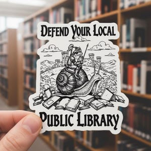 May include: A white sticker with the text "Defend Your Local Public Library." The sticker features a whimsical illustration of a frog knight riding a snail, with a castle and books in the background.