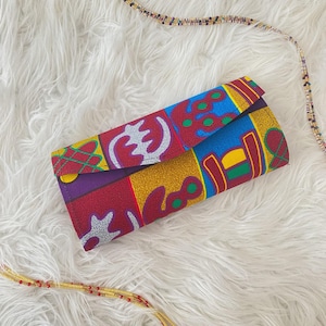 May include: A colorful clutch with a geometric pattern in red, blue, yellow, and purple. The clutch is rectangular and folded over, with a flap closure. The fabric has a glittery texture. A beaded necklace is visible in the background.