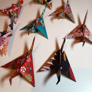Könnte beinhalten: Eine Sammlung von Origami-Kranichen in verschiedenen Farben und Mustern. Die Papierkraniche sind aus gemustertem Papier mit floralen, geometrischen und abstrakten Designs gefaltet. Die Kraniche sind auf einer weißen Oberfläche angeordnet.