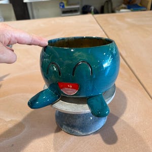 May include: A teal ceramic planter with a smiling face design. The planter has a round body with two arm-like extensions and a red mouth. The interior of the planter is a darker shade of teal.