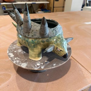 May include: A ceramic sculpture of a creature with a spiky back and a fierce expression. The sculpture is primarily green and gray, with a dark interior. It sits on a round, speckled base, showcasing a unique, handcrafted design.