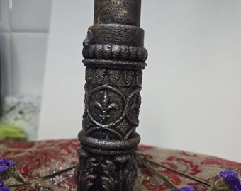 May include: A dark brown pillar candle with ornate detailing, including fleur-de-lis patterns and textured bands. The candle has a rough, textured surface and a white wick. The base is a matching dark brown color. Purple flowers are in the foreground.