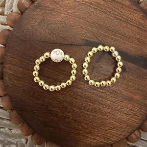 May include: Two gold beaded rings, one with a white bead featuring a cross design. The rings are displayed on a dark brown wooden surface with a decorative border. The rings are likely for the fingers.