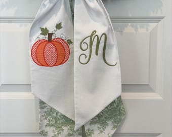 May include: White fabric door hanger with a pumpkin applique and the letter "M" embroidered in green. The bottom of the hanger has a green and white toile pattern. The hanger is tied to a green wreath.