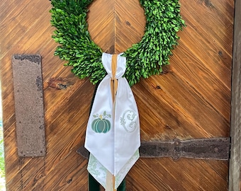 May include: A vibrant green wreath made of boxwood, adorned with a white fabric sash. The sash features a green pumpkin and floral embroidery, with gold and green ribbons. The wreath hangs on a rustic wooden door.
