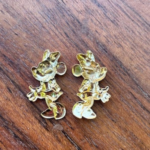 May include: Two gold-colored Minnie Mouse-shaped earrings. Each earring features a detailed design of the iconic character, including her signature bow and dress. The earrings are displayed on a wooden surface.
