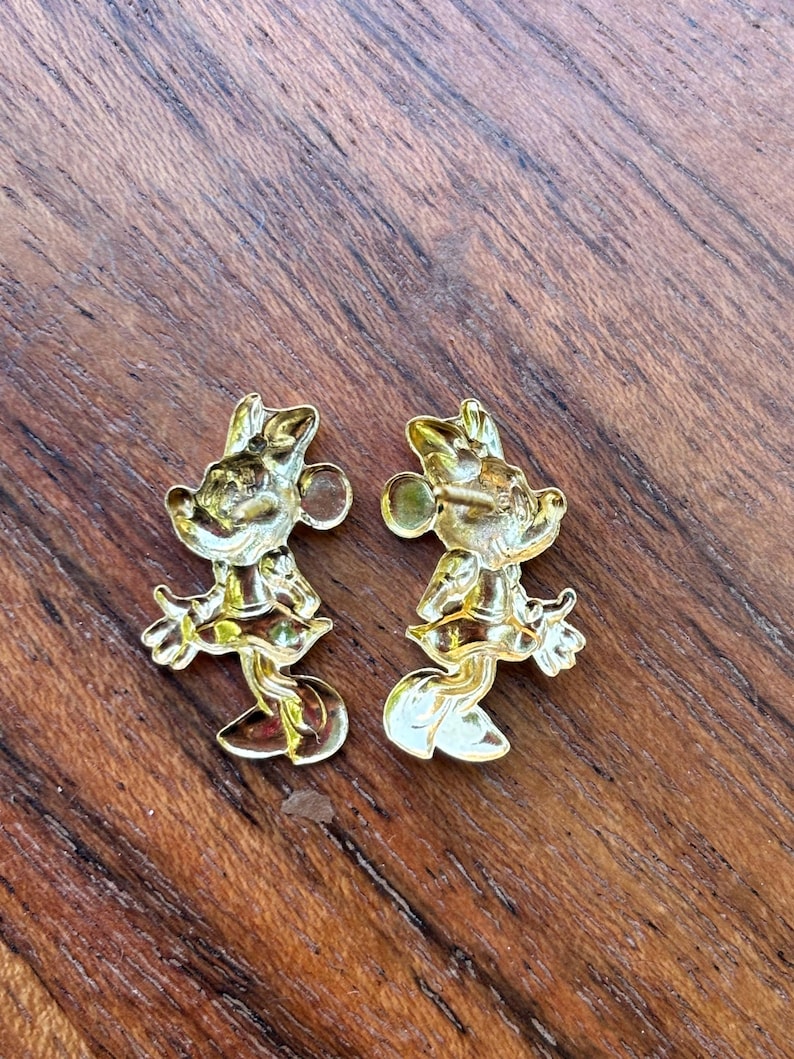 May include: Two gold-colored Minnie Mouse-shaped earrings. Each earring features a detailed design of the iconic character, including her signature bow and dress. The earrings are displayed on a wooden surface.