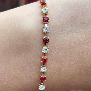 May include: A gold-toned bracelet featuring alternating red and clear square-cut gemstones. The bracelet is shown on a person's wrist, with the gemstones catching the light. The bracelet is a piece of jewelry.