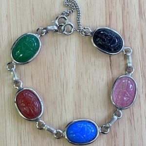 May include: A silver-toned link bracelet featuring five oval-shaped, carved scarab-style stones in green, black, pink, blue, and red. The bracelet includes a silver-toned metal chain with a circular spring ring clasp and a short extender chain.