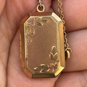 May include: A gold-toned rectangular locket with floral and leaf designs. The locket is suspended from a delicate gold chain. The locket has an octagonal shape with a hinged opening. The floral details are in a darker gold color.