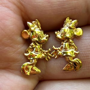 May include: A pair of gold-colored Minnie Mouse stud earrings. Each earring features a detailed, three-dimensional design of Minnie Mouse in her classic pose, complete with a bow and dress. The earrings are small and likely made of metal.
