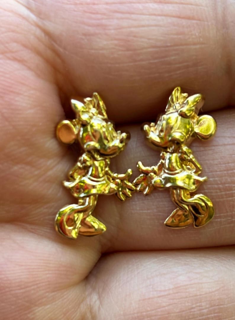 May include: A pair of gold-colored Minnie Mouse stud earrings. Each earring features a detailed, three-dimensional design of Minnie Mouse in her classic pose, complete with a bow and dress. The earrings are small and likely made of metal.