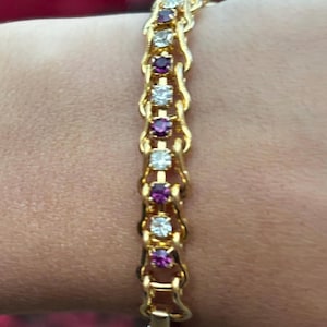 May include: A gold-toned bracelet featuring an alternating pattern of round, clear white stones and round, deep magenta-colored stones. The stones are set within a decorative, interlocking gold-toned metal link chain that secures with a metal clasp.