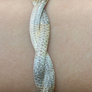 May include: A gold-colored bracelet with a braided design. The bracelet is made of a textured material and is wrapped around a wrist. The bracelet's design is a series of interwoven, flat, textured bands.