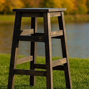 May include: A dark brown wooden bar stool with a square seat and a ladder-style frame. The stool is made of stained wood and has a simple, rustic design. It is set on a grassy area with a lake and trees in the background.