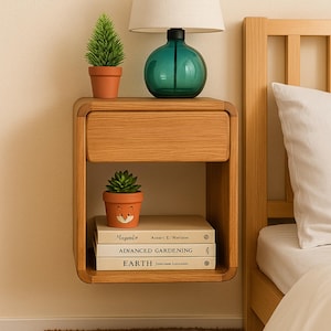 Puede incluir: Una mesita de noche flotante de madera con un cajón, que contiene una lámpara de cristal verde, pequeñas plantas en macetas y libros. Los libros se titulan "Maple", "Advanced Gardening" y "Earth". La mesita de noche está junto a una cama con una almohada blanca.