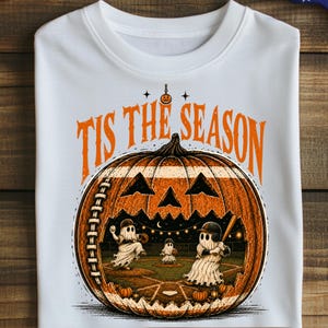 Puede incluir: Una camiseta blanca con una gran calabaza naranja tallada como una pelota de béisbol, con el texto 'TIS THE SEASON' encima. Dentro de la calabaza, tres fantasmas vestidos de beisbolistas están en un campo bajo luces nocturnas.