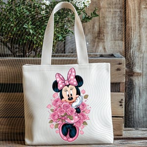 May include: A cream-colored tote bag featuring a Minnie Mouse graphic. Minnie is depicted in a pink polka-dot dress and bow, surrounded by pink roses. The bag has two cream-colored handles and is set against a wooden background.