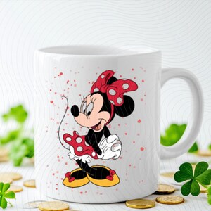 May include: White ceramic mug featuring a colorful illustration of Minnie Mouse. Minnie is depicted in her classic outfit: a red dress with white polka dots, a matching bow, and yellow shoes. The mug has a curved handle.