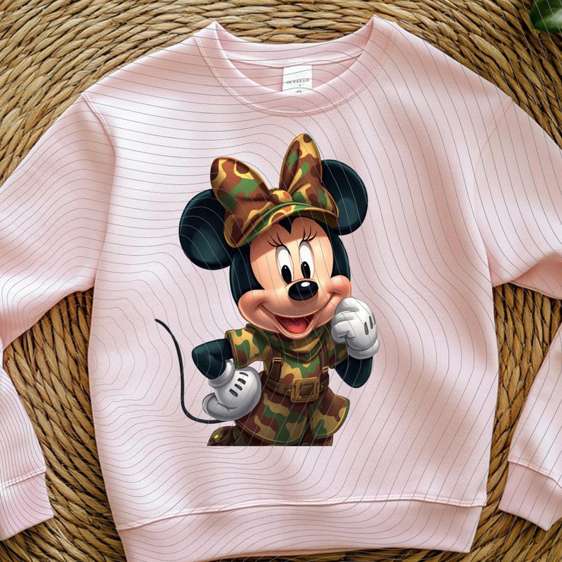 Pode incluir: Uma camisola de gola redonda rosa claro com uma impress&atilde;o gr&aacute;fica de uma personagem de rato de desenho animado a usar um conjunto com padr&atilde;o de camuflagem e um la&ccedil;o a condizer. A camisola tem um padr&atilde;o subtil de linhas onduladas.
