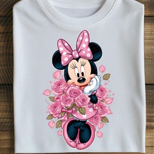 May include: White t-shirt featuring a cartoon Minnie Mouse design. Minnie Mouse is wearing a pink polka dot dress and bow, surrounded by pink roses. The design is centered on the shirt.