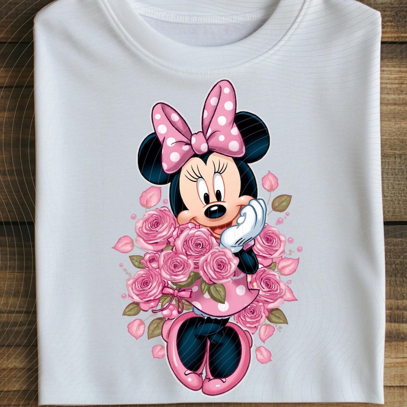 May include: White t-shirt featuring a cartoon Minnie Mouse design. Minnie Mouse is wearing a pink polka dot dress and bow, surrounded by pink roses. The design is centered on the shirt.