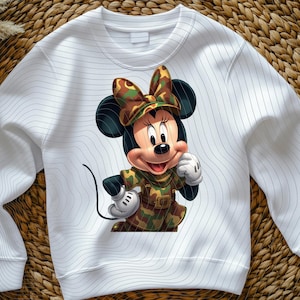 Pode incluir: Uma camisola branca de manga comprida com o gr&aacute;fico de um rato de desenho animado a usar um conjunto com estampado de camuflagem e um la&ccedil;o a condizer. O tecido branco apresenta um padr&atilde;o subtil de linhas horizontais finas e onduladas.