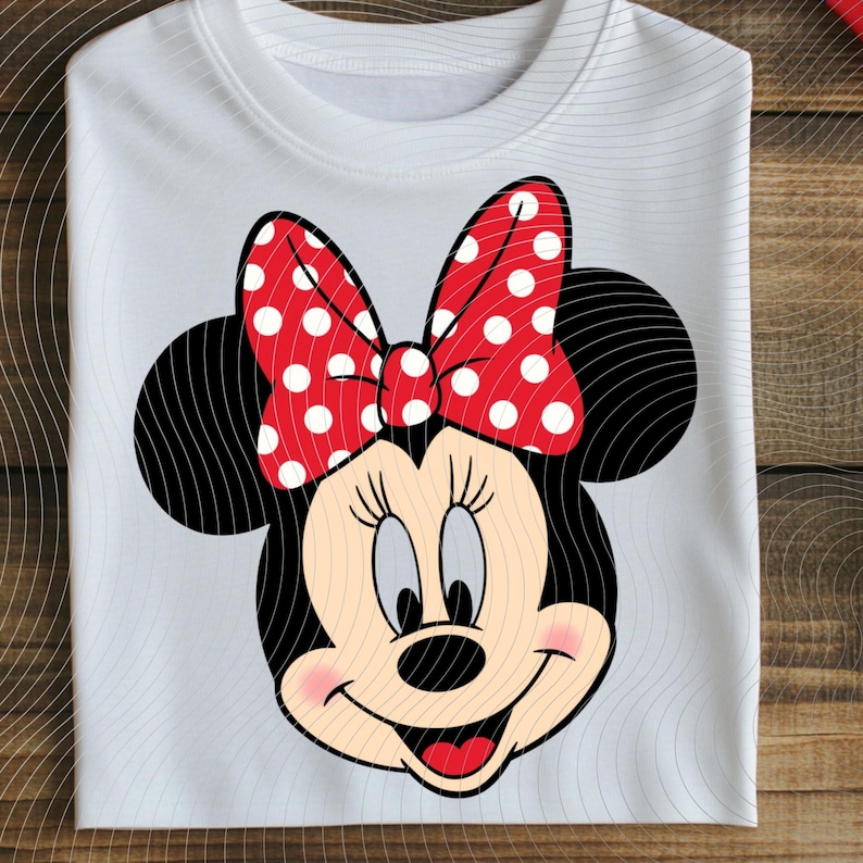 Op de afbeelding: Wit T-shirt met een Minnie Mouse-afbeelding. Het ontwerp toont zwarte oren, een rode strik met witte stippen en een lachend gezicht met roze wangen. Het T-shirt is op een houten oppervlak gevouwen.