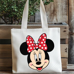 Op de afbeelding: Een witte canvas draagtas met een cartoonafbeelding van Minnie Mouse. De afbeelding toont Minnie's gezicht met zwarte oren, een rode strik met witte stippen en een glimlachende uitdrukking. De tas heeft twee witte handvatten.