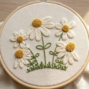 May include: A circular wooden embroidery hoop containing a piece of off-white fabric. The fabric features hand-stitched embroidery of four daisy flowers with white petals and textured yellow centers, connected by green stems and small leaves.