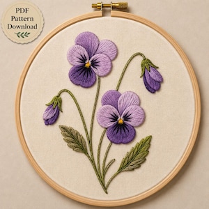 May include: An embroidery hoop containing a fabric panel with a hand-stitched design of two purple pansy flowers, two closed buds, and green leaves. The flowers feature shades of light and dark purple with yellow centers and dark purple details.