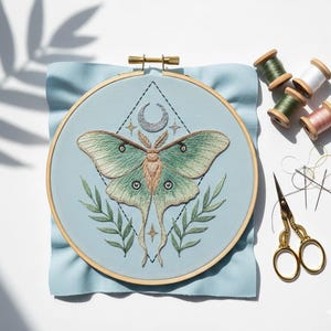 May include: An embroidered moth design on light blue fabric, set in a wooden hoop. The moth is green and tan, with a geometric design and crescent moon. Includes spools of thread, needles, and gold scissors.