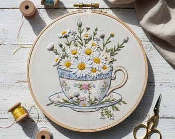 Puede incluir: Arte floral bordado en un aro de madera. El diseño presenta una taza de té rebosante de margaritas blancas con centros amarillos. La taza y el platillo están decorados con patrones florales azules y rosas. También se ven carretes de hilo de bordar y tijeras doradas.