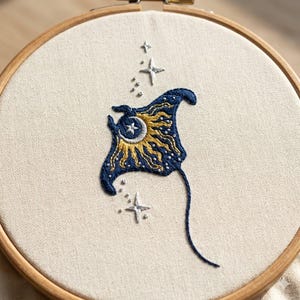 May include: Embroidered art piece featuring a navy blue manta ray with a gold sun and moon design. The artwork includes embroidered stars and is set in a wooden embroidery hoop. The background is a light beige fabric.