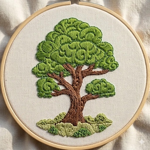 May include: Hand-stitched embroidery art of a tree in a wooden hoop. The tree features a brown trunk and branches with lush green leaves. The base of the tree has green shrubbery, all on a cream-colored fabric.