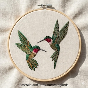 Op de afbeelding: Geborduurd kunstwerk met twee kolibries in vlucht. De vogels hebben groene en bruine vleugels, rode kelen en lange zwarte snavels. De tekst "Emerald and Ruby Hummingbirds" staat onderaan. De borduursel zit in een houten ring.