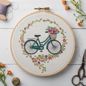 May include: An embroidery hoop featuring a teal bicycle with a basket of colorful flowers. The bicycle is surrounded by a floral wreath with red, orange, and purple flowers. The embroidery is set against a white fabric background.